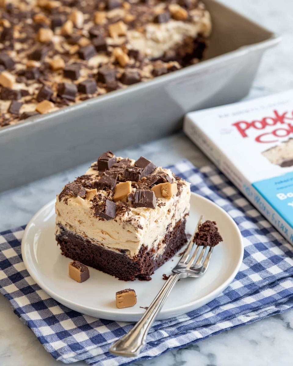 The image shows a thick square piece of chocolate cake on a white plate resting on a white marbled surface with a blue cloth underneath. The cake has two layers: a dark brown moist chocolate cake at the bottom and a light brown creamy frosting on top sprinkled with many small pieces of peanut butter cups that add texture and color. Next to the plate, a fork holds a piece of cake ready to be eaten. Behind, there is a baking pan filled with more of the same cake, with a layer of light brown frosting and a generous topping of chopped peanut butter cups. The photo taken with an iphone --ar 4:5 --v 7