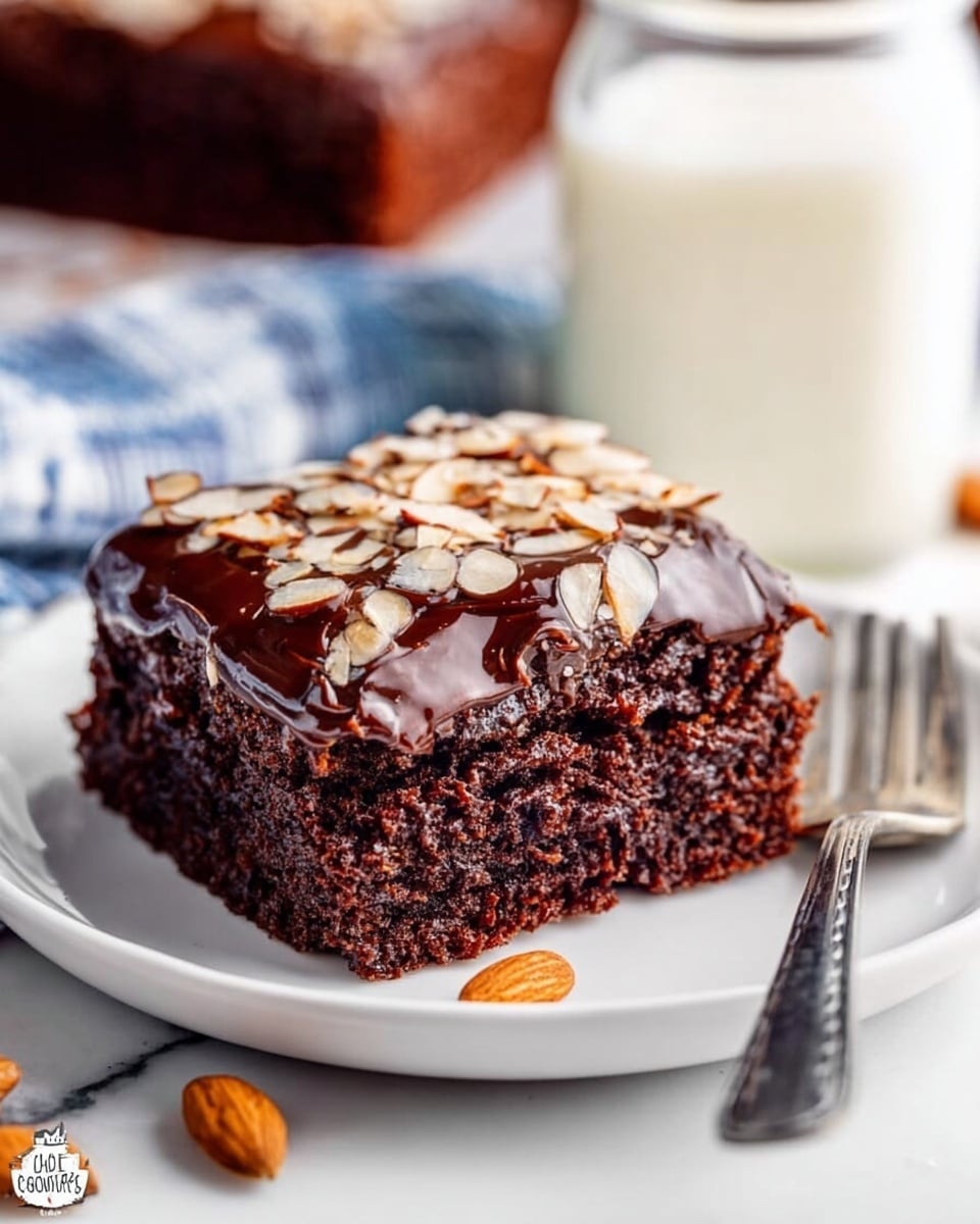 A square piece of dark brown chocolate cake is on a white plate. The cake has two layers: the bottom layer is soft and moist chocolate cake, while the top layer is shiny chocolate frosting. Thin, light tan almond slices are scattered all over the frosting. There are a few almond slices around the plate. A silver fork is placed behind the cake on the plate. In the blurred background, there is a glass jar with a white liquid, likely milk, sitting on a white marbled surface. Photo taken with an iphone --ar 4:5 --v 7