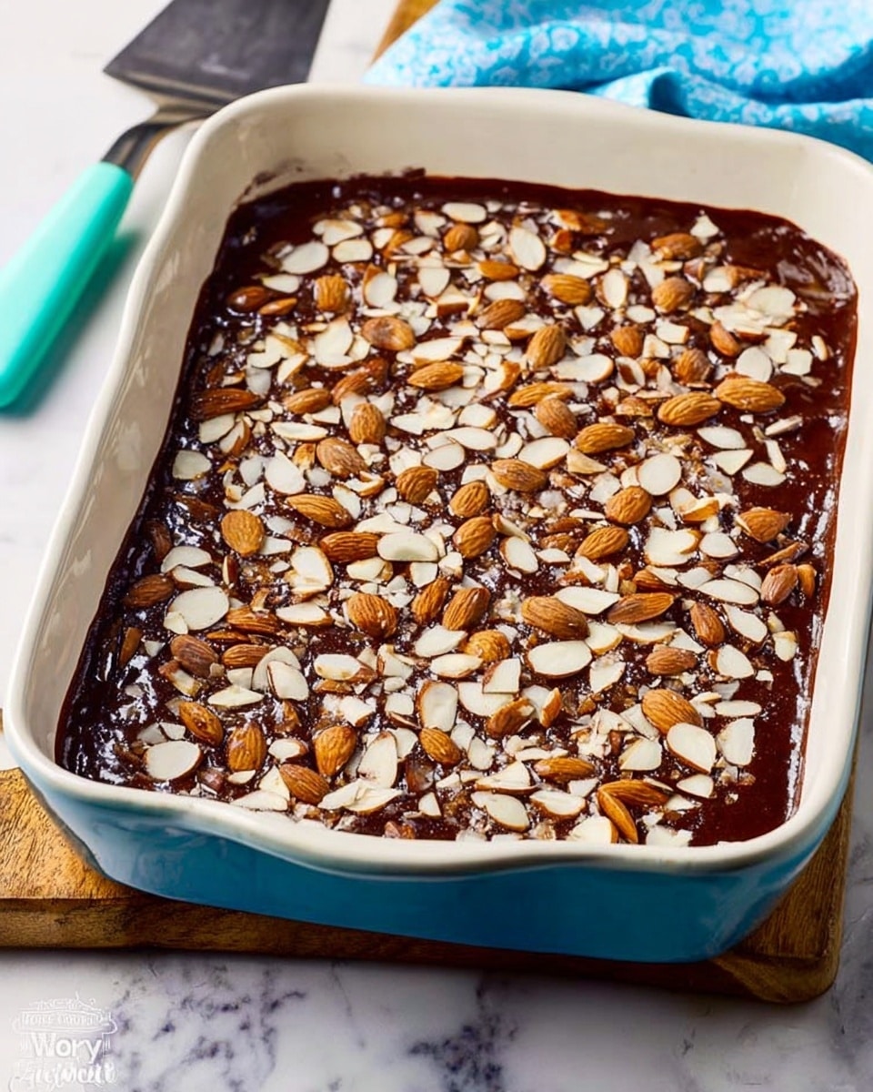 A rectangular white baking dish filled with a thick layer of dark brown chocolate topping, spread evenly across the surface. The top is covered with a generous amount of sliced almonds, scattered all over with different sizes and light brown toasted edges, giving a crunchy texture. The dish sits on a white marbled surface, next to a metal spatula with a turquoise handle. A light blue cloth is blurred in the background. Photo taken with an iphone --ar 4:5 --v 7