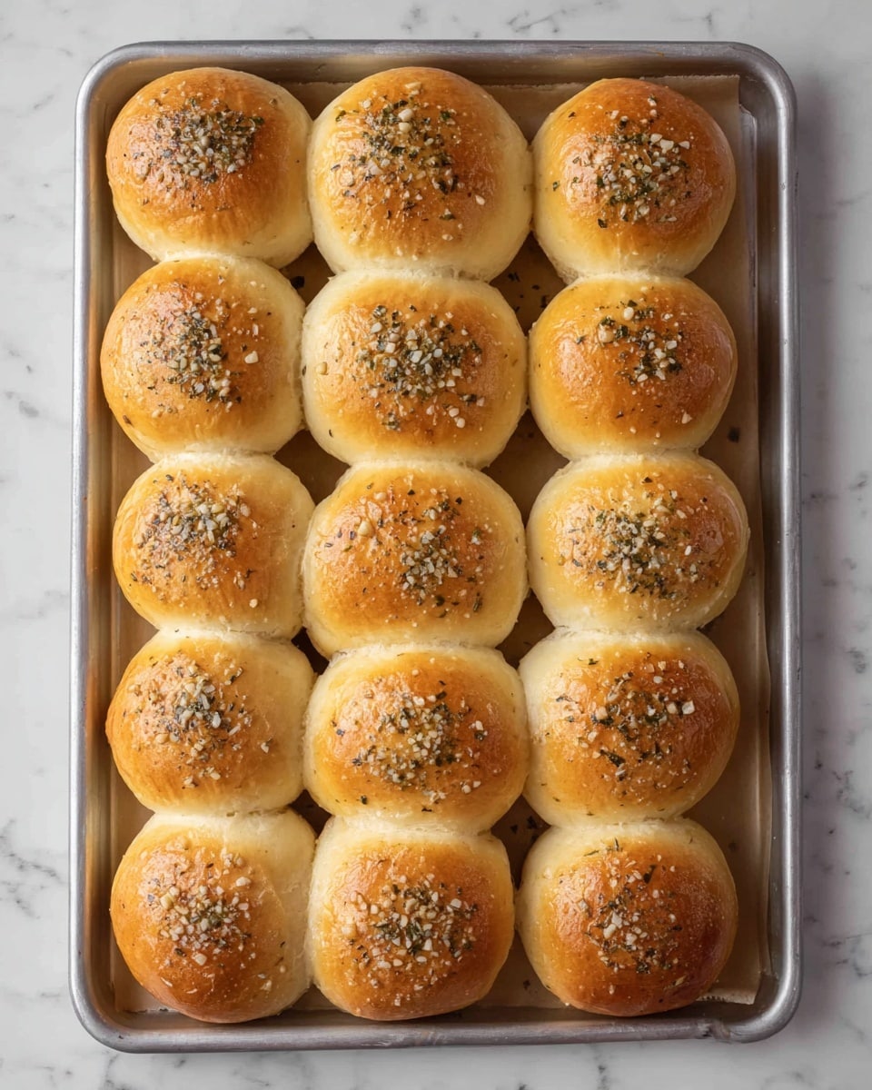 A tray of 16 round golden brown bread rolls is neatly arranged in four rows and four columns on a baking sheet lined with parchment paper. Each roll has a shiny, smooth top sprinkled with a mix of chopped garlic and herbs that add texture with small bits spread unevenly. The rolls are close together, showing soft, fluffy sides where they touch. The baking sheet is placed on a white marbled surface. photo taken with an iphone --ar 4:5 --v 7