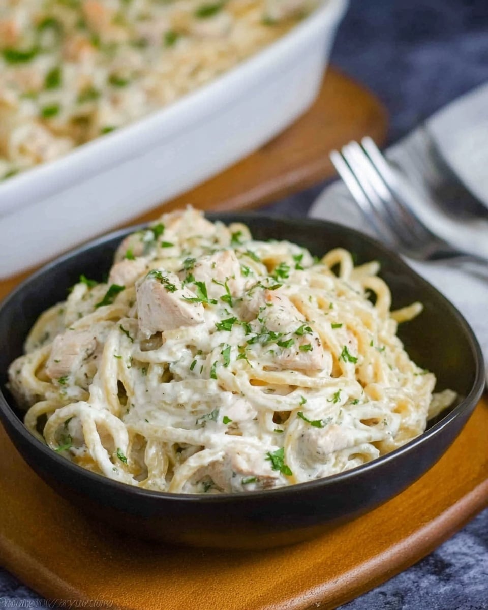 A dark bowl filled with a creamy white pasta dish layered with tender white chicken pieces and finely chopped green herbs sprinkled on top. The pasta strands are coated in a rich white sauce that looks thick and smooth. The bowl is placed on a wooden surface with a silver fork lying nearby on a white marbled background. In the background, there is a white baking dish filled with more of the creamy pasta, slightly blurred. The photo taken with an iphone --ar 4:5 --v 7