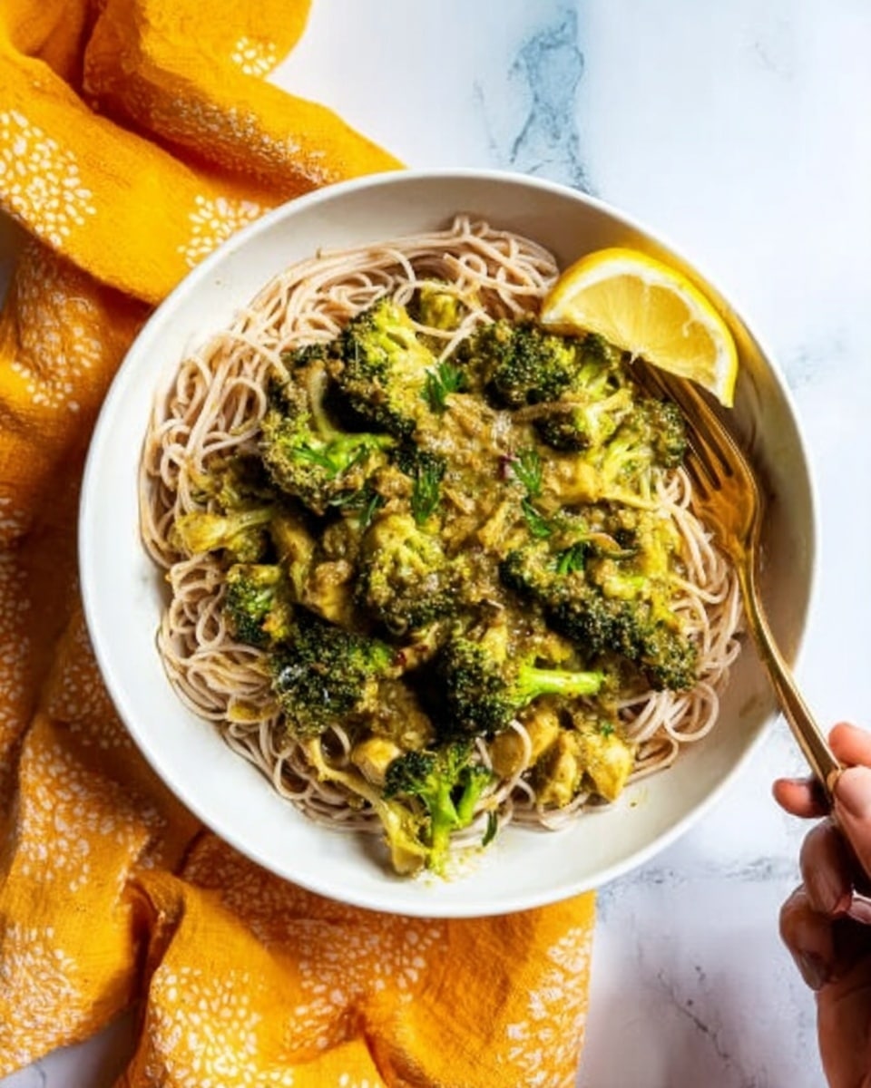 A white plate holds a base layer of light beige noodles with a slightly rough texture, topped with bright green roasted broccoli florets that show some browned, crispy edges. A thin twist of yellow lemon zest is sprinkled over the broccoli, and a lemon wedge sits on the right side of the plate. The plate is set on a white marbled surface, with a golden fork placed below the front edge. In the background, there is a second white plate with the same noodle and broccoli dish, and a yellow and blue cloth adds color behind the plates. Photo taken with an iphone --ar 4:5 --v 7