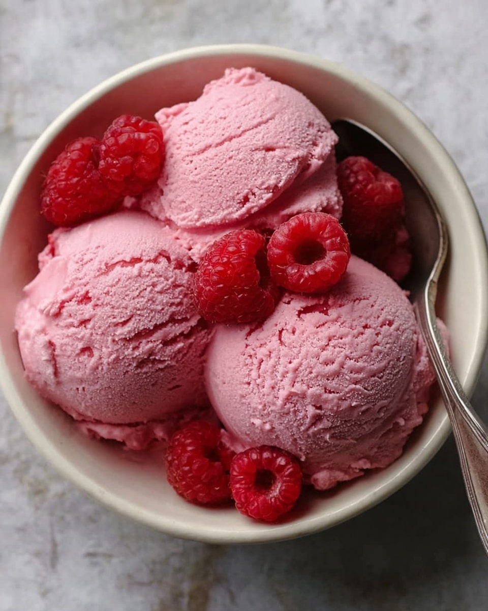 The image shows a white bowl filled with three large scoops of pink ice cream with a smooth but slightly textured surface. On top of the ice cream, there are several bright red raspberries scattered, adding contrast and freshness to the dish. A silver spoon lies inside the bowl on the right side, partially visible. The bowl sits on a surface with a white marbled texture. photo taken with an iphone --ar 4:5 --v 7