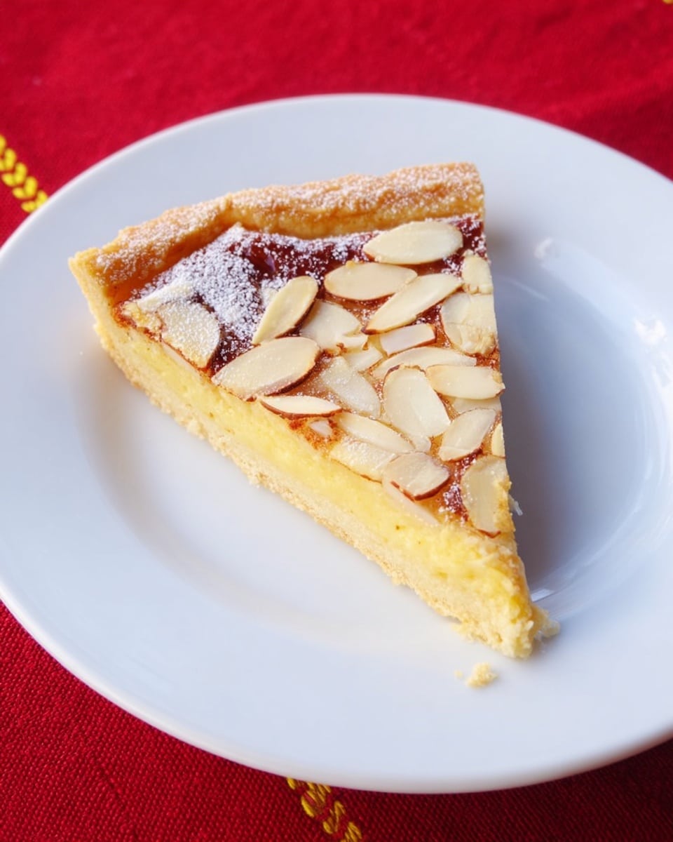 The image shows a single slice of almond tart placed in the center of a white plate. The tart has three visible layers: a golden-brown crust at the bottom, a thick light yellow almond filling in the middle, and a top layer sprinkled with thin sliced almonds and a light dusting of sugar. The crust looks crisp and slightly flaky, while the almond filling appears smooth with a soft texture. The white plate sits on a red fabric with a yellow stitch line visible on the left edge. Photo taken with an iphone --ar 4:5 --v 7