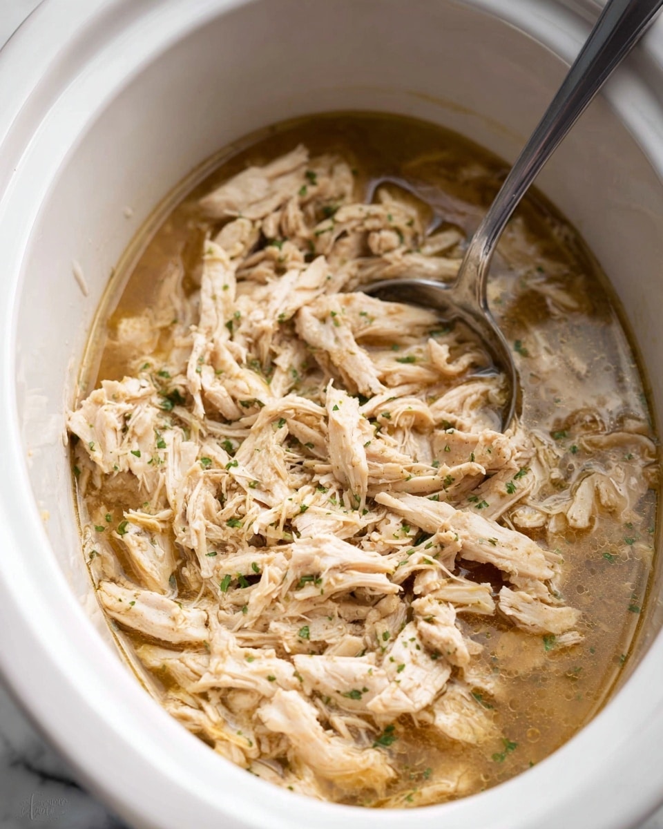 This image shows a close-up of a white slow cooker filled with shredded chicken in a light brown broth, with small green herb flecks scattered on top. The shredded chicken pieces are soft, pale beige, and layered evenly across the cooker. A large metal spoon is partially resting inside the cooker, mixing the chicken and broth. The cooker sits on a white marbled surface. Photo taken with an iphone --ar 4:5 --v 7