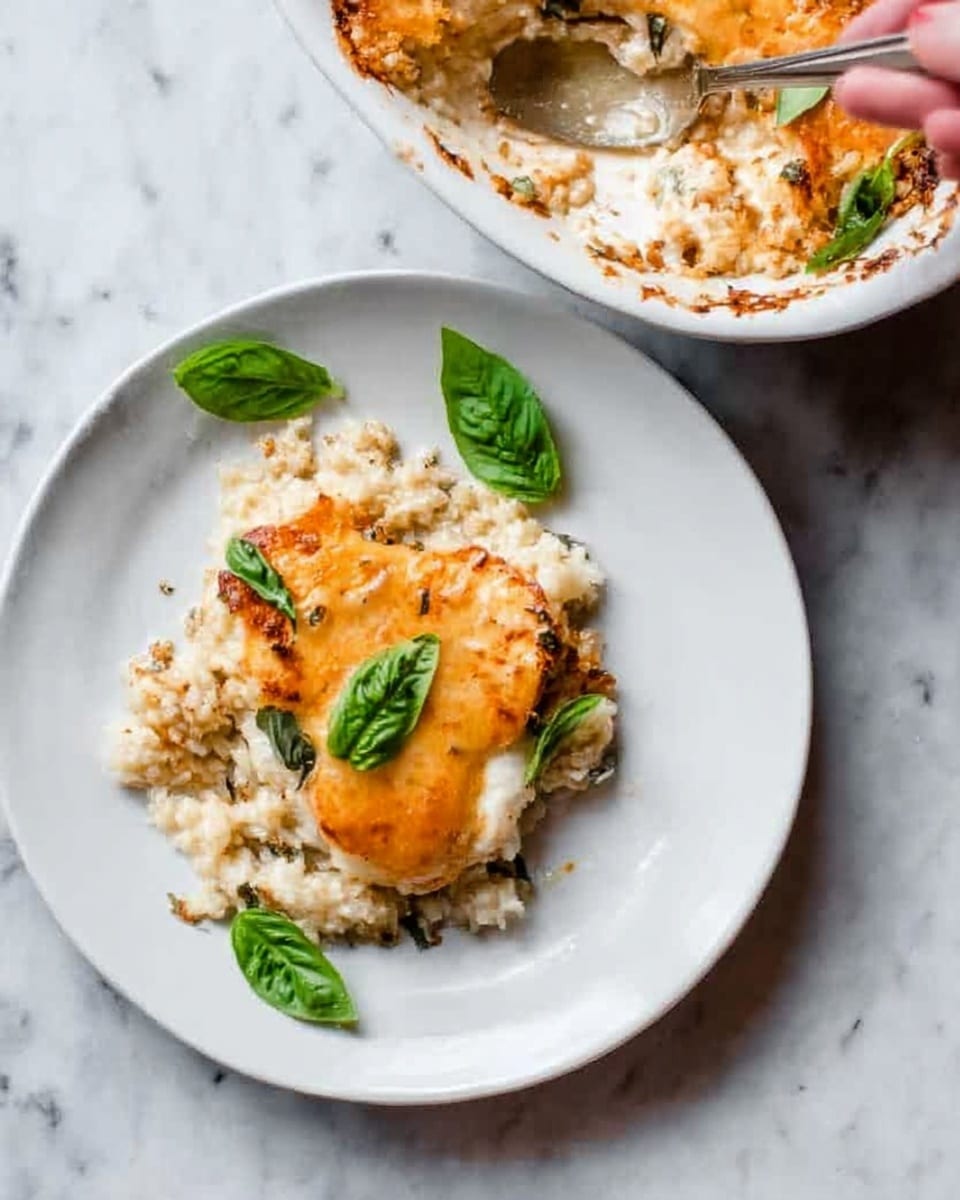 A white plate holds a serving of a baked cheesy dish with a golden-orange melted cheese layer on top. Underneath the cheese, there is a creamy, light texture that looks like a rice or grain base, slightly browned around the edges. Small bright green basil leaves are placed on top of the cheese and around the dish, adding a fresh contrast. The plate rests on a white marbled surface, and a white bowl containing more of the same dish is partially visible above, with a woman's hand spooning some out. Photo taken with an iphone --ar 4:5 --v 7