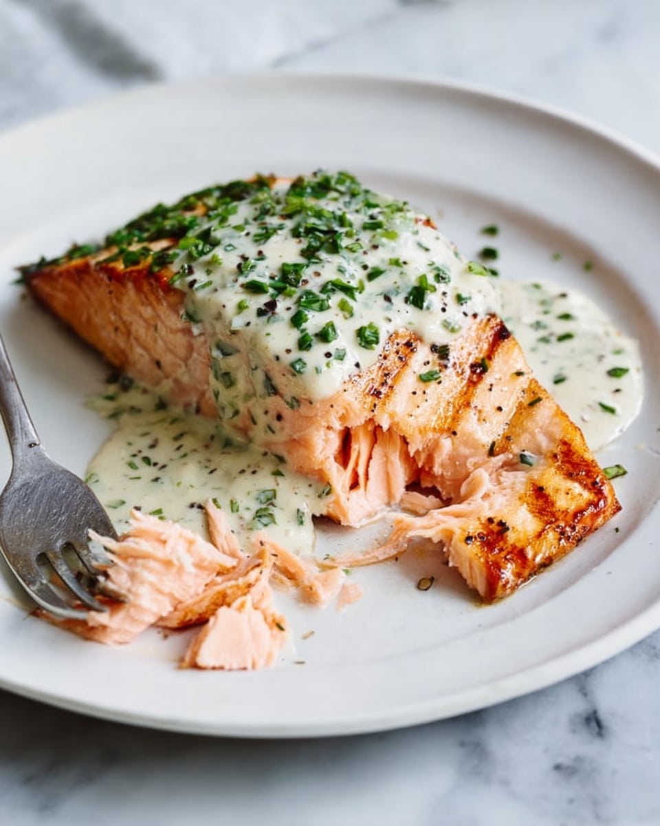 A white round plate holds a large piece of cooked salmon with a golden grilled texture on top and a soft pink inside. The salmon is topped with a thick creamy white sauce mixed with green herbs and specks of black pepper. Small pieces of salmon are flaked near the bottom left of the fish, with a silver fork resting beside them on the plate. The plate sits on a white marbled surface. photo taken with an iphone --ar 4:5 --v 7
