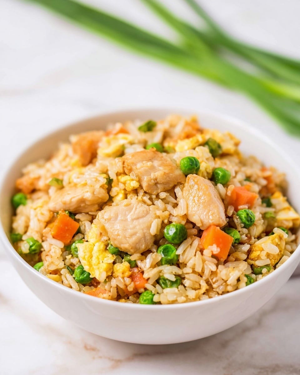 A white bowl filled with fried rice that has several layers of ingredients mixed together. The base layer is light brown rice grains, followed by small green peas scattered evenly. Bright orange carrot cubes are spread throughout along with light yellow scrambled egg pieces. Chunky pieces of light golden chicken sit on top, giving the dish varied texture and color. The white bowl is placed on a white marbled surface with some green onion stalks blurred in the background. photo taken with an iphone --ar 4:5 --v 7