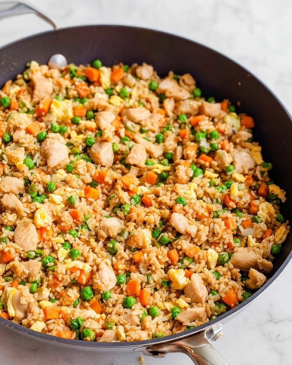 The image shows a pan filled with fried rice mixed with many small, light brown chicken pieces, bright green peas, orange carrot cubes, and scrambled egg bits scattered throughout. The rice is golden and evenly spread, creating a textured layer. The pan is round, dark, and deep with a shiny metallic handle, placed on a white marbled surface. The colors are warm and inviting, highlighting the mix of ingredients in a simple, homely style photo taken with an iphone --ar 4:5 --v 7