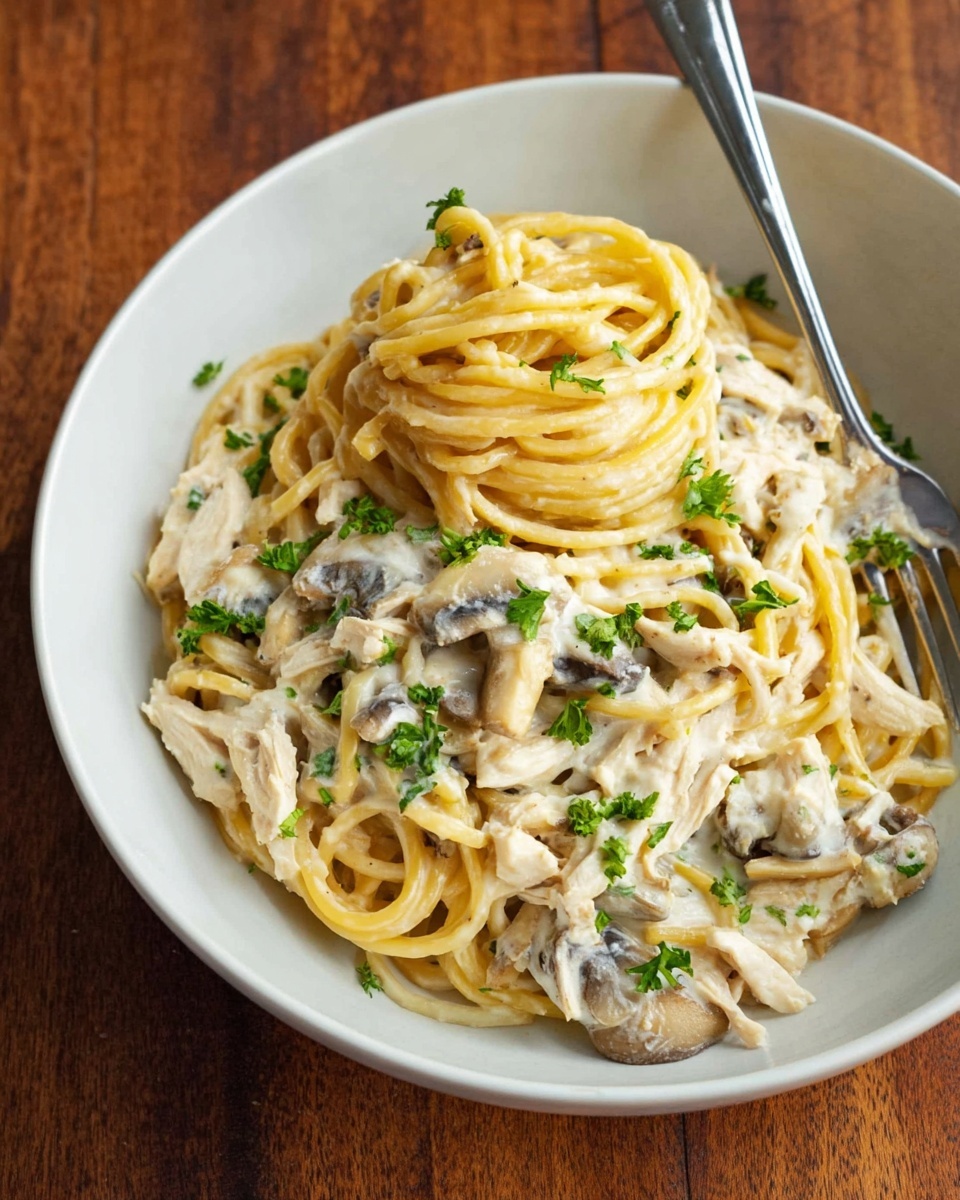 A white bowl holds a creamy pasta dish with two main layers: thick spaghetti noodles on top that are a pale yellow with a smooth texture, and a creamy white sauce underneath mixed with pieces of light brown mushrooms and white chicken strips. Small green parsley leaves are sprinkled over the dish for color. A silver fork rests on the right side inside the bowl. The bowl sits on a wooden surface. photo taken with an iphone --ar 4:5 --v 7
