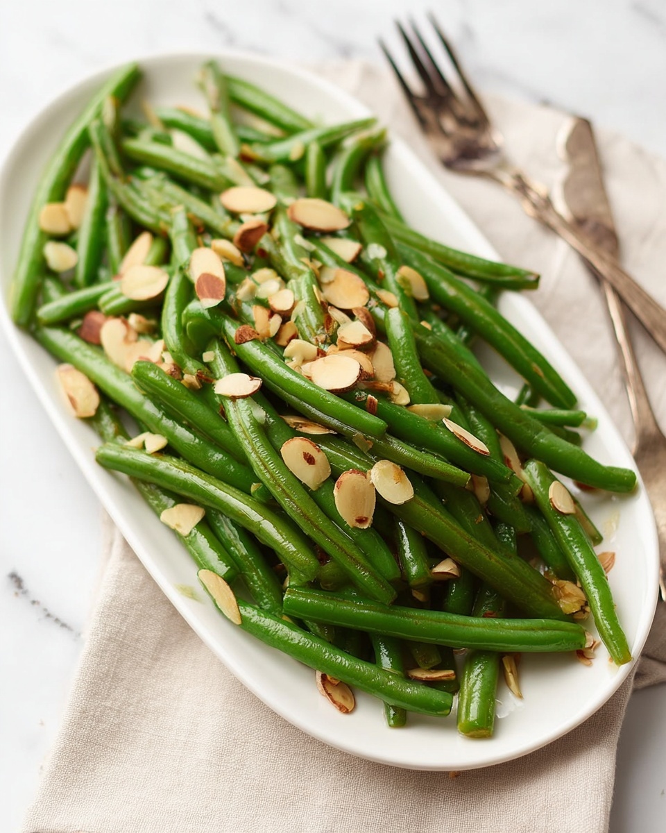 A white rectangular plate holds a single layer of bright green cooked green beans spread evenly across the plate. On top, scattered thinly sliced toasted almond pieces add a light brown color and crunchy texture contrast. The plate is placed on a white marbled surface, and near it, part of a fork and a beige cloth napkin are visible in soft focus. The image is bright and clear, showing the fresh and simple look of the green bean dish. photo taken with an iphone --ar 4:5 --v 7