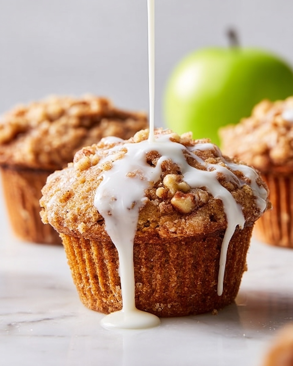 A close-up of a golden-brown muffin with a crumbly, textured top layer that has small nut pieces scattered on it. White icing is being poured over the muffin from above, flowing in smooth, thick lines that drip down the sides. In the blurred background, there are two more similar muffins and a green apple, all placed on a white marbled surface. The overall lighting is soft and natural, highlighting the warm tones of the muffin and the smooth whiteness of the icing. Photo taken with an iphone --ar 4:5 --v 7