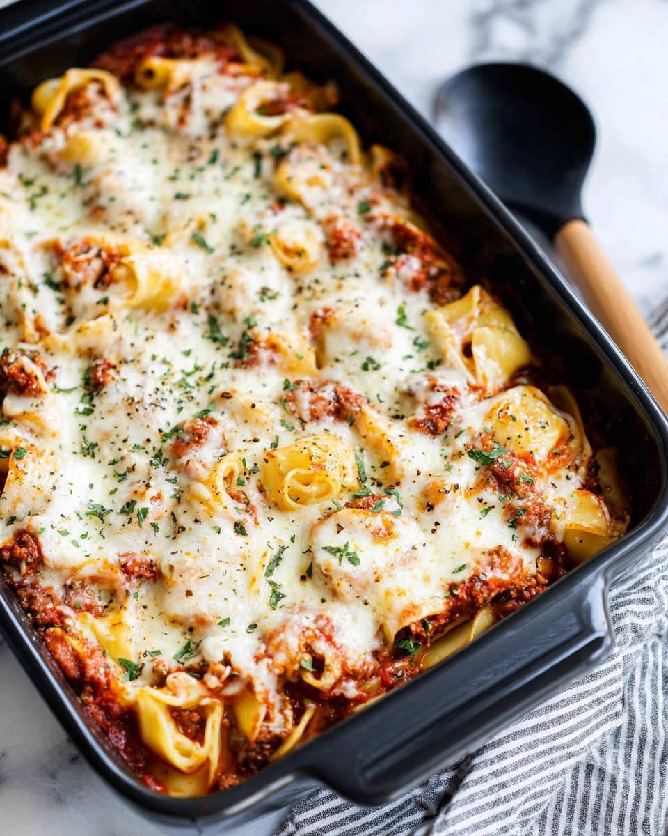 A close-up view of a black rectangular baking dish filled with a layered pasta bake topped with melted creamy white cheese sprinkled with green herbs and black pepper. The top layer has thick, pale yellow pasta rings interspersed with chunky, rich red tomato sauce mixed with browned meat pieces. The edges of the dish show a slight crisping of the sauce and cheese. A wooden-handled black spoon rests on the top right edge of the dish. The dish sits on a white marbled surface with a striped cloth partially visible on the bottom right. photo taken with an iphone --ar 4:5 --v 7