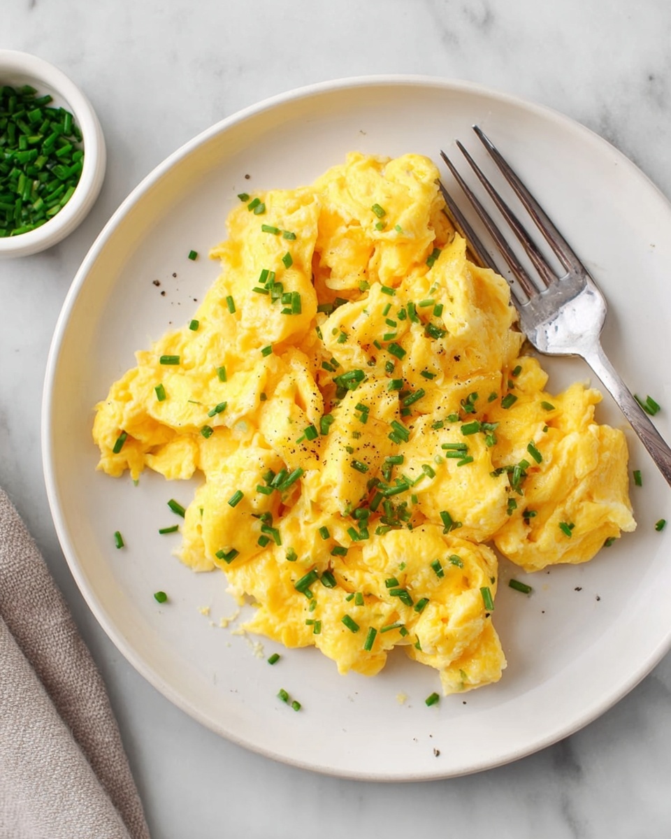 The image shows a close-up of a white plate filled with bright yellow scrambled eggs that look soft and fluffy. The eggs have small curvy layers and a creamy texture, topped with small pieces of chopped green chives scattered evenly across the top. A silver fork is placed on the right side of the plate, resting on the white marbled surface. The light is soft, making the eggs look fresh and warm, photo taken with an iphone --ar 4:5 --v 7