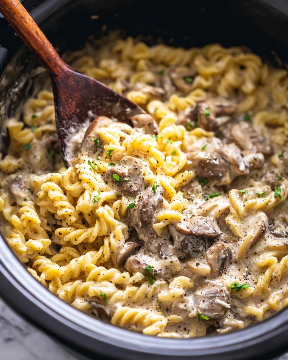 The image shows a black slow cooker filled with creamy pasta and mushroom stew. The bottom layer is smooth, light beige sauce with specks of black pepper, mixed with tender pieces of brown mushrooms and chunks of meat. On top, there are yellow spiral pasta pieces coated in the sauce, scattered evenly. Small green herb sprigs rest on the surface, adding a fresh touch. A wooden spoon with a textured, dark handle is inside the cooker, gently stirring the dish. The cooker is placed on a white marbled surface. Photo taken with an iphone --ar 4:5 --v 7