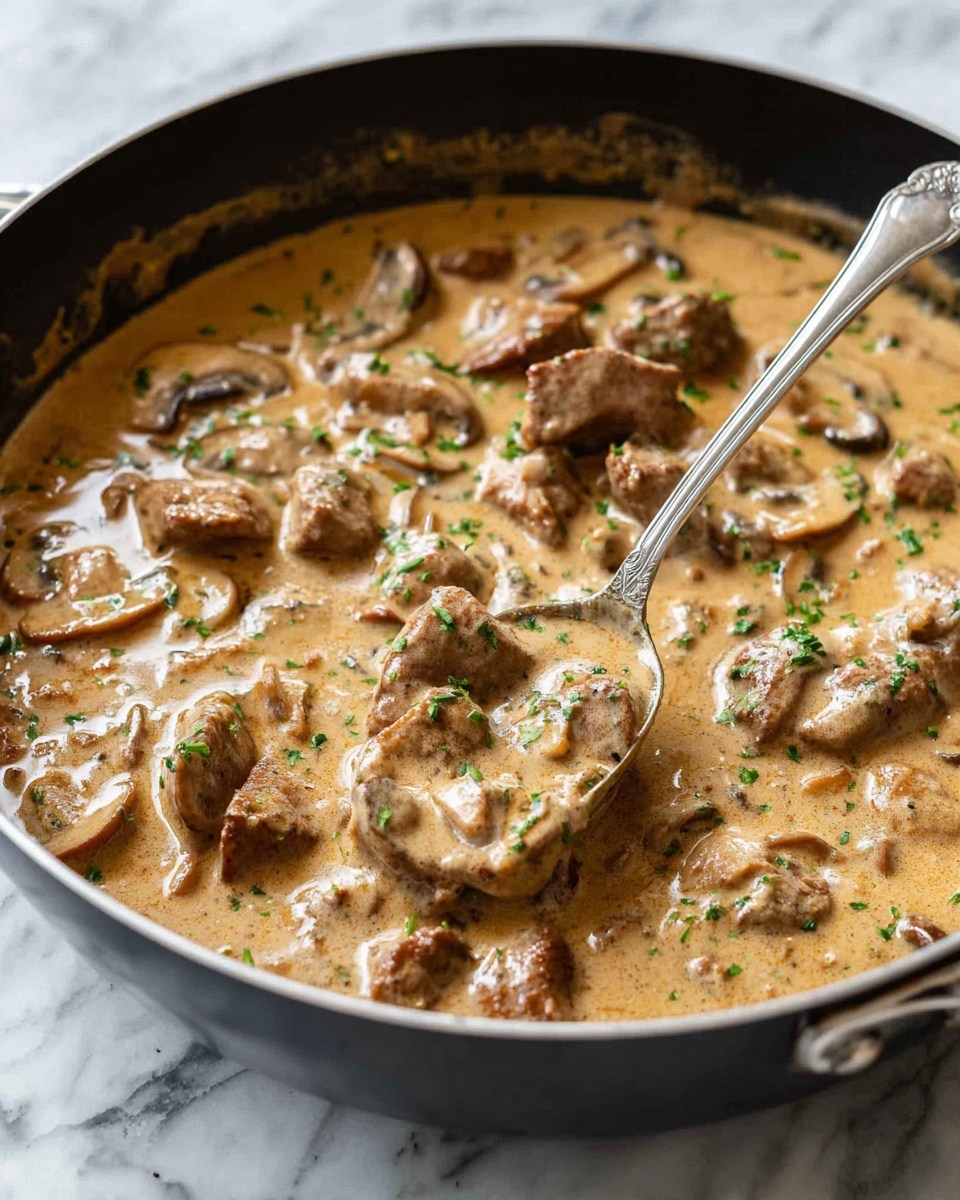 A close-up view of a creamy mushroom and meat dish in a white bowl, filled with a light brown sauce that has a smooth, thick texture. The dish contains several layers of sliced mushrooms, both whole and halved, mixed with brown pieces of tender meat, all coated evenly in the sauce. Small green bits, likely chopped herbs, are sprinkled on top, adding a touch of fresh color. A silver spoon is partially submerged on the right side of the bowl, resting inside. The bowl is placed on a white marbled surface. Photo taken with an iphone --ar 4:5 --v 7