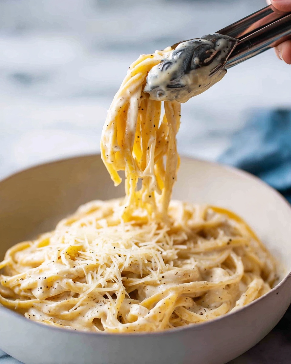 A white bowl filled with creamy pasta noodles, coated in a smooth light beige sauce with specks of black pepper, topped with a generous layer of finely grated pale yellow cheese. A pair of metal tongs held by a woman's hand lifts a twisted bundle of noodles from the bowl, showing the thick sauce stretching slightly. The bowl is placed on a white marbled surface with blurred white and blue background. photo taken with an iphone --ar 4:5 --v 7