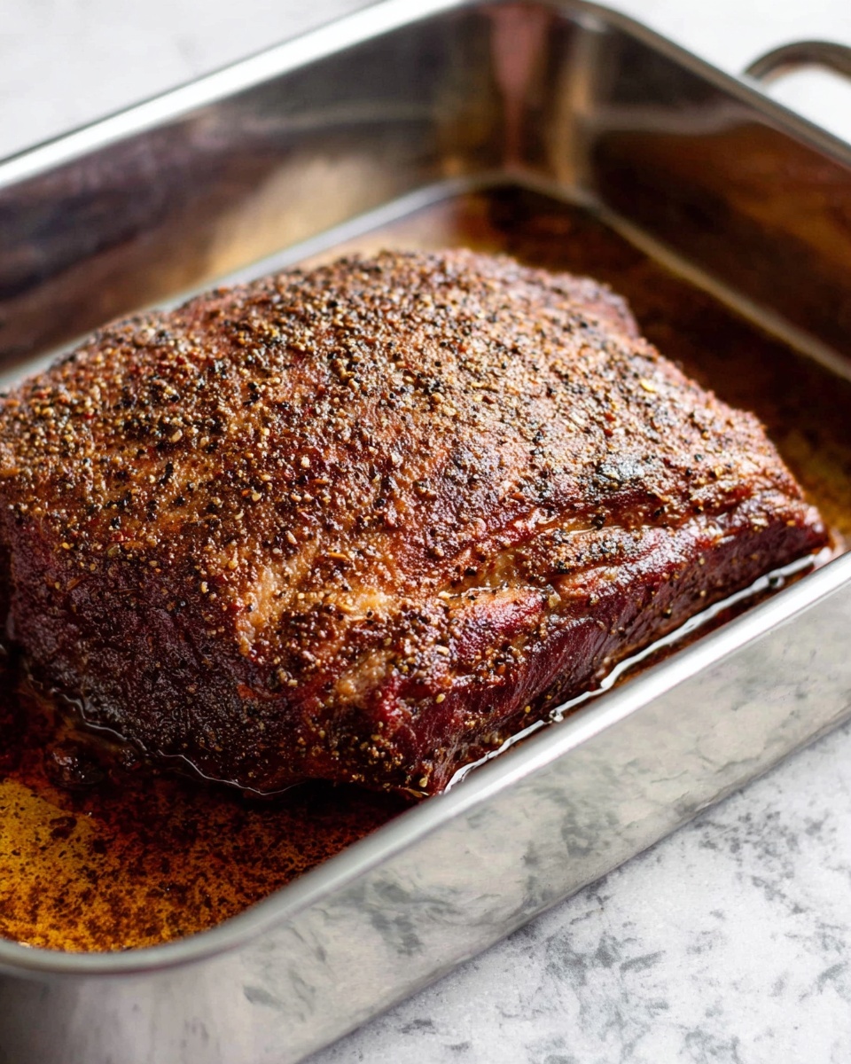 A close-up view of a single large piece of cooked meat resting in a shiny metal roasting pan. The meat has a thick, textured outer layer that is dark brown with visible specks of black pepper and spices, showing a slightly crispy and seasoned crust. The pan contains a small pool of juices around the meat, reflecting light softly. The background surface surrounding the pan is a white marbled texture. photo taken with an iphone --ar 4:5 --v 7