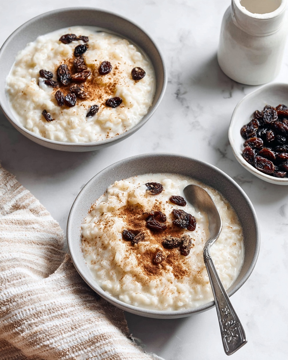 A silver pan filled with creamy rice pudding that has a smooth, thick texture and visible cooked rice grains, dotted with dark raisins throughout. A spoon rests inside the pan, partly submerged in the pudding. Around the pan, there are small bowls containing ground cinnamon and more raisins, all placed on a white marbled surface. A striped beige and black cloth lies near the pan’s handle, adding a soft texture to the scene. photo taken with an iphone --ar 4:5 --v 7