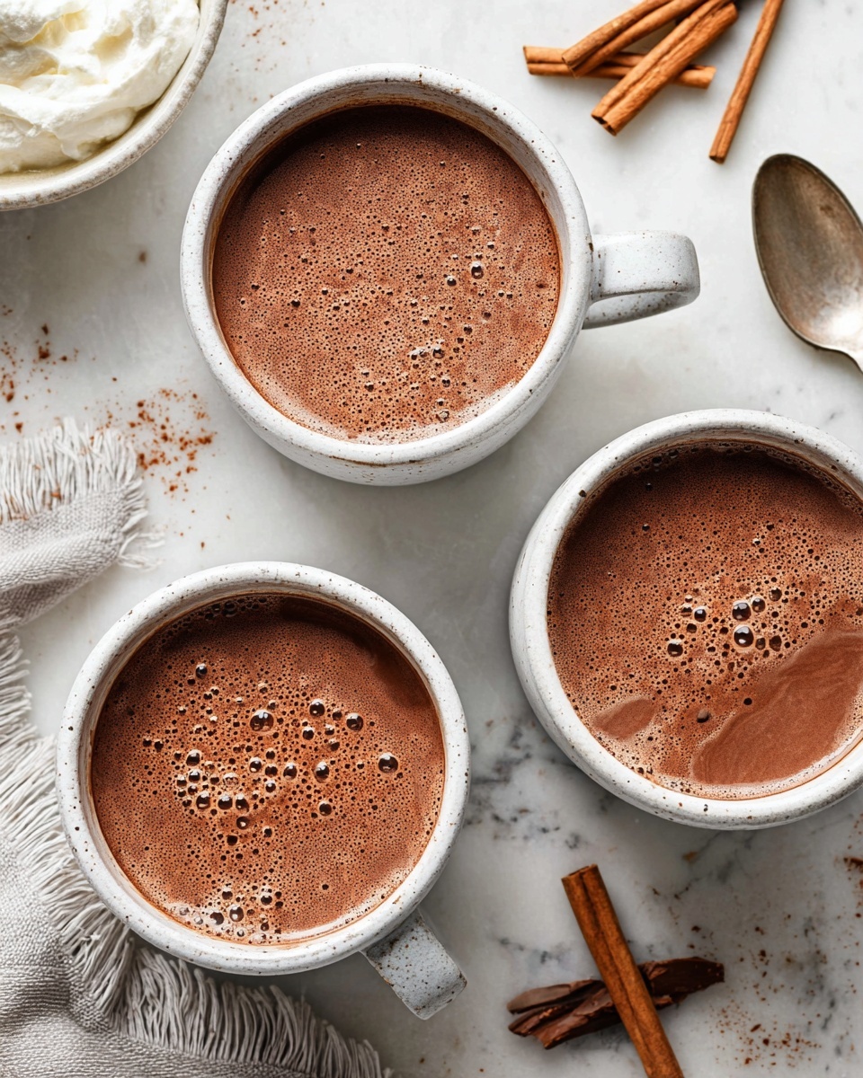 A close-up image shows a cup filled with hot chocolate, with a light brown frothy top layer covered in many tiny bubbles. The cup is white with a light brown base and a thick handle on the right side. To the left, there is a white bowl holding thick white cream with a spoon resting inside it. In the background on the white marbled surface, there are some cinnamon sticks lying next to the cup. A white cloth with some frayed edges is partially visible in the lower right corner. The lighting is soft and natural, giving a cozy feeling. photo taken with an iphone --ar 4:5 --v 7