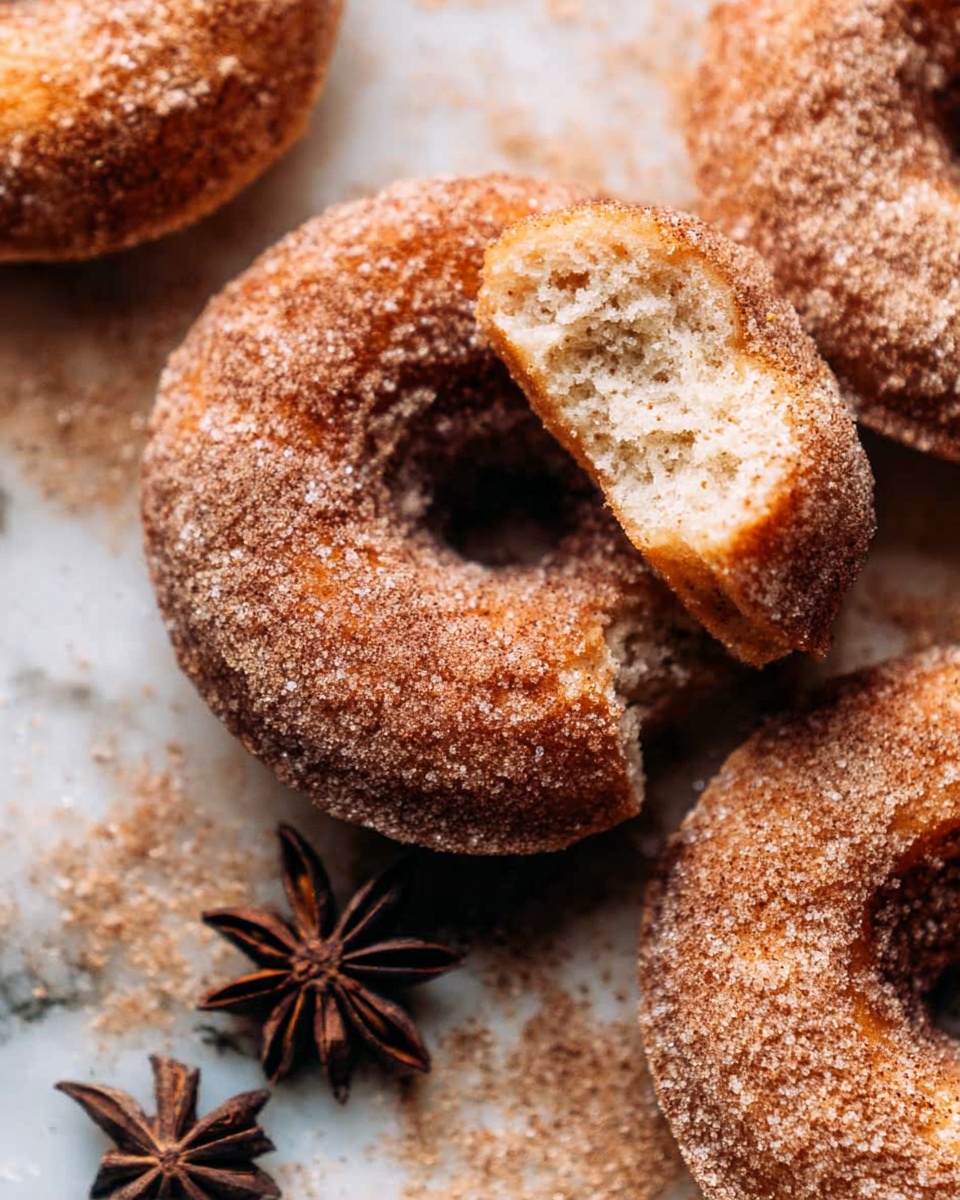 The image shows three cinnamon sugar donuts placed on a white marbled surface dusted with granulated sugar. One donut is broken in half, revealing a soft, fluffy inside with a light beige color. The donuts are coated evenly with a sparkling layer of cinnamon sugar, giving them a rough texture in warm brown and golden tones. Two star anise spices are placed near the donuts, adding a dark brown star shape contrast. The close-up view captures the cozy, sweet feel of the treats. Photo taken with an iphone --ar 4:5 --v 7