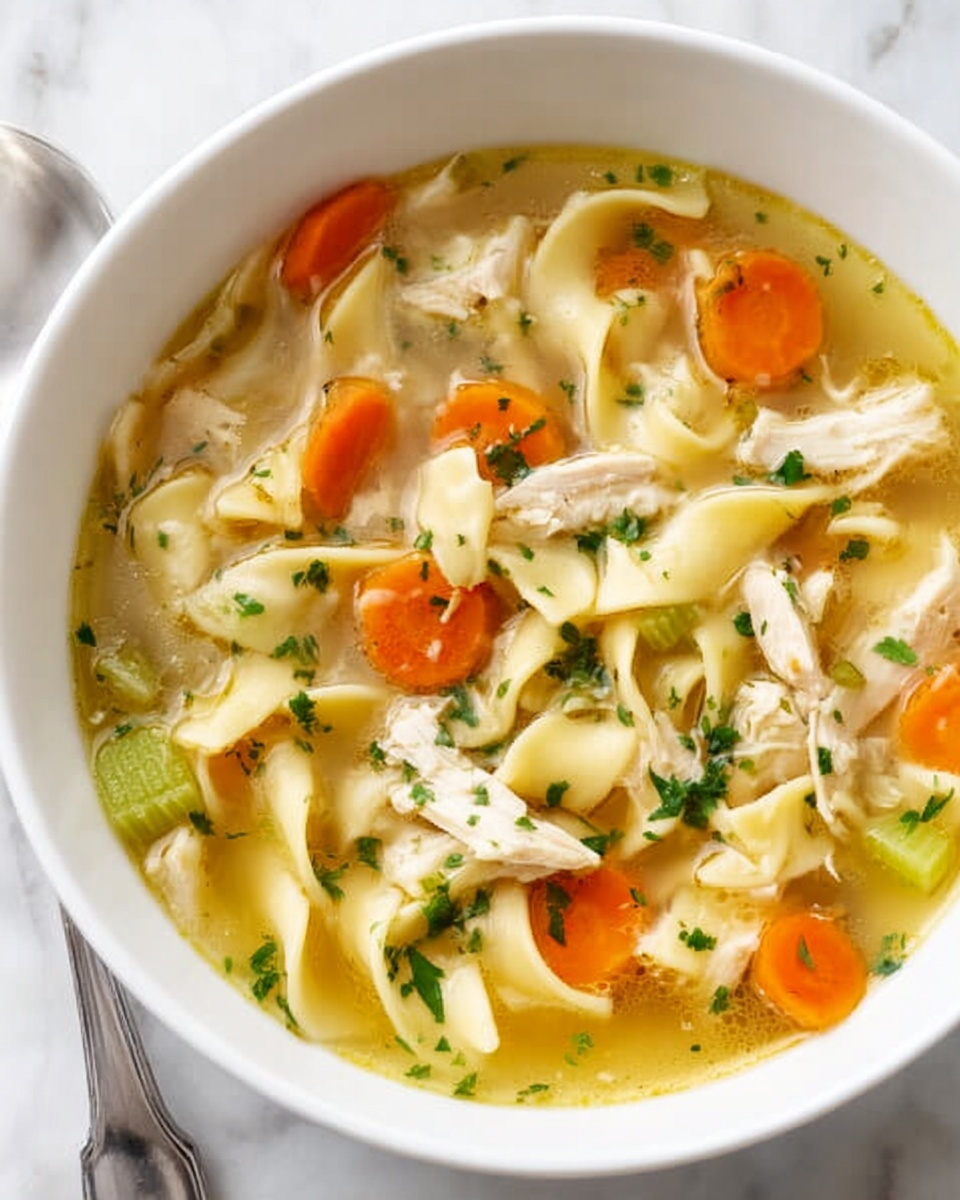 A white bowl filled with chicken noodle soup resting on a white marbled surface. The soup has clear golden broth with visible small pieces of white cooked chicken, thick flat yellow noodles, bright orange carrot slices, and pale green celery pieces. Chopped green herbs sprinkled on top add a fresh touch. A silver spoon is placed near the bowl. photo taken with an iphone --ar 4:5 --v 7