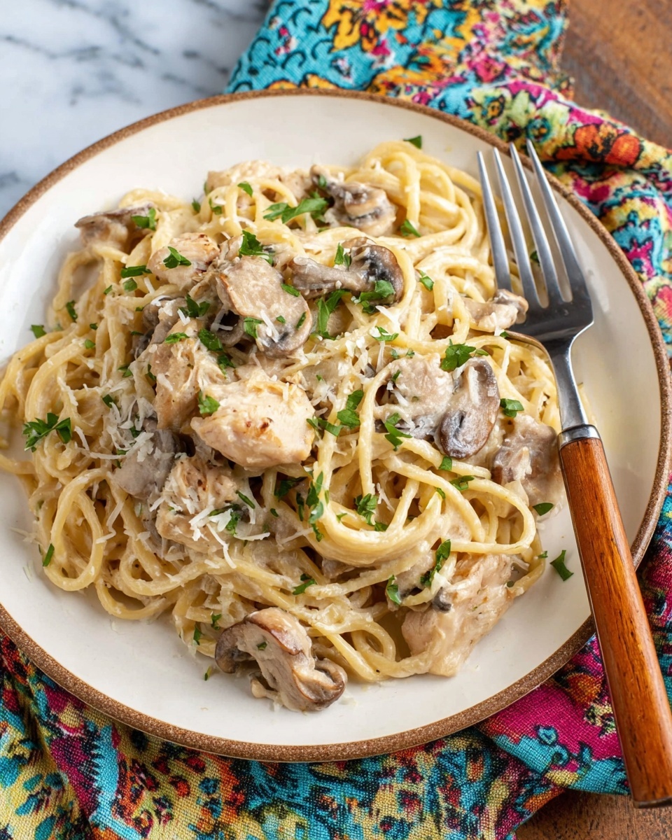 A close-up view of creamy pasta with three main layers: the bottom layer is light yellow cooked spaghetti mixed with small chunks of pale white chicken pieces; the middle layer has browned, tender-looking mushroom slices scattered evenly; the top layer is sprinkled with finely shredded white cheese and small bright green parsley leaves. This is all inside a round metal pan, resting on a colorful floral cloth over a wooden table, with a white marbled texture visible at the edges. The dish looks creamy and inviting. photo taken with an iphone --ar 4:5 --v 7