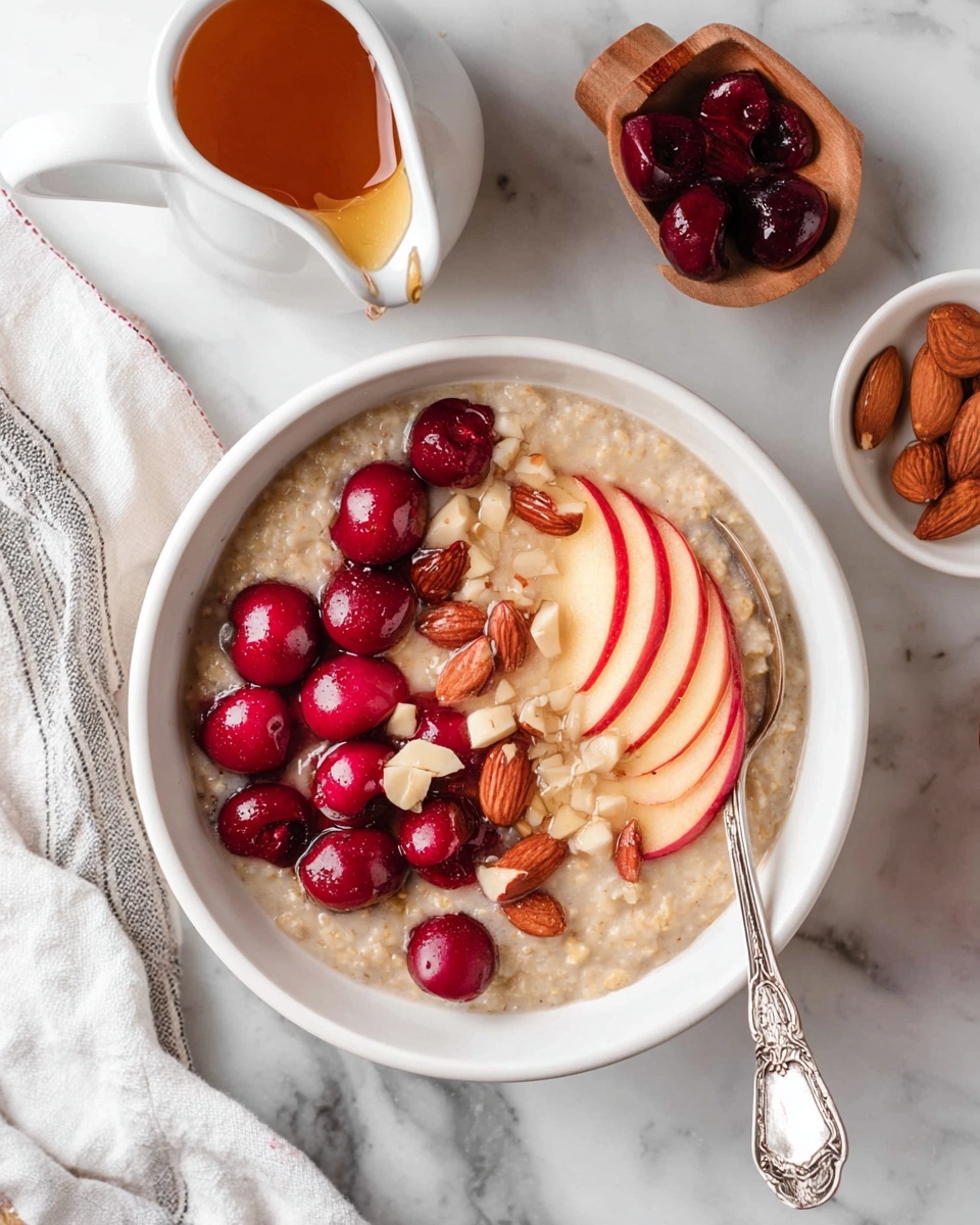 Two white bowls of porridge sit on a white marbled surface. The bowl on the top right has a creamy porridge base topped with a cluster of red cherries in the center, thin pale pink apple slices arranged in a fan shape to the right, and small chopped almonds scattered around. A silver spoon is placed in this bowl. The bowl on the bottom left has a creamy porridge base topped with a dollop of white yogurt in the center, fresh dark blueberries around the yogurt, white coconut flakes, and thin orange peel curls sprinkled on top. A silver spoon is lying nearby. Around the bowls are a cup of dark tea with a tea bag, a small white pitcher of honey or syrup, a small bowl of coconut flakes, a small wooden scoop filled with chopped almonds, a wooden cutting board with a sliced red apple and a knife, and a beige cloth with black stripes. Photo taken with an iphone --ar 4:5 --v 7