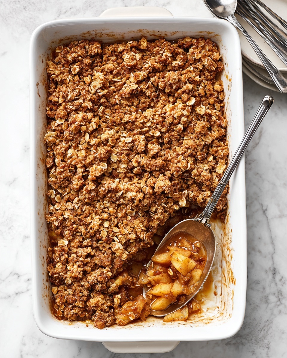 This is a white rectangular baking dish filled with a layered apple crumble. The bottom layer is soft, golden-brown cooked apple pieces soaked in a syrupy juice, seen where a portion has been scooped out. The top layer is a thick, uneven crumble made with oats and brown sugar, textured and golden with hints of cinnamon. A large metal spoon rests inside the dish, partly submerged in the apple layer, positioned at the bottom right corner. The dish sits on a white marbled surface with part of another white bowl visible on the top right and two silver forks on the left. photo taken with an iphone --ar 4:5 --v 7
