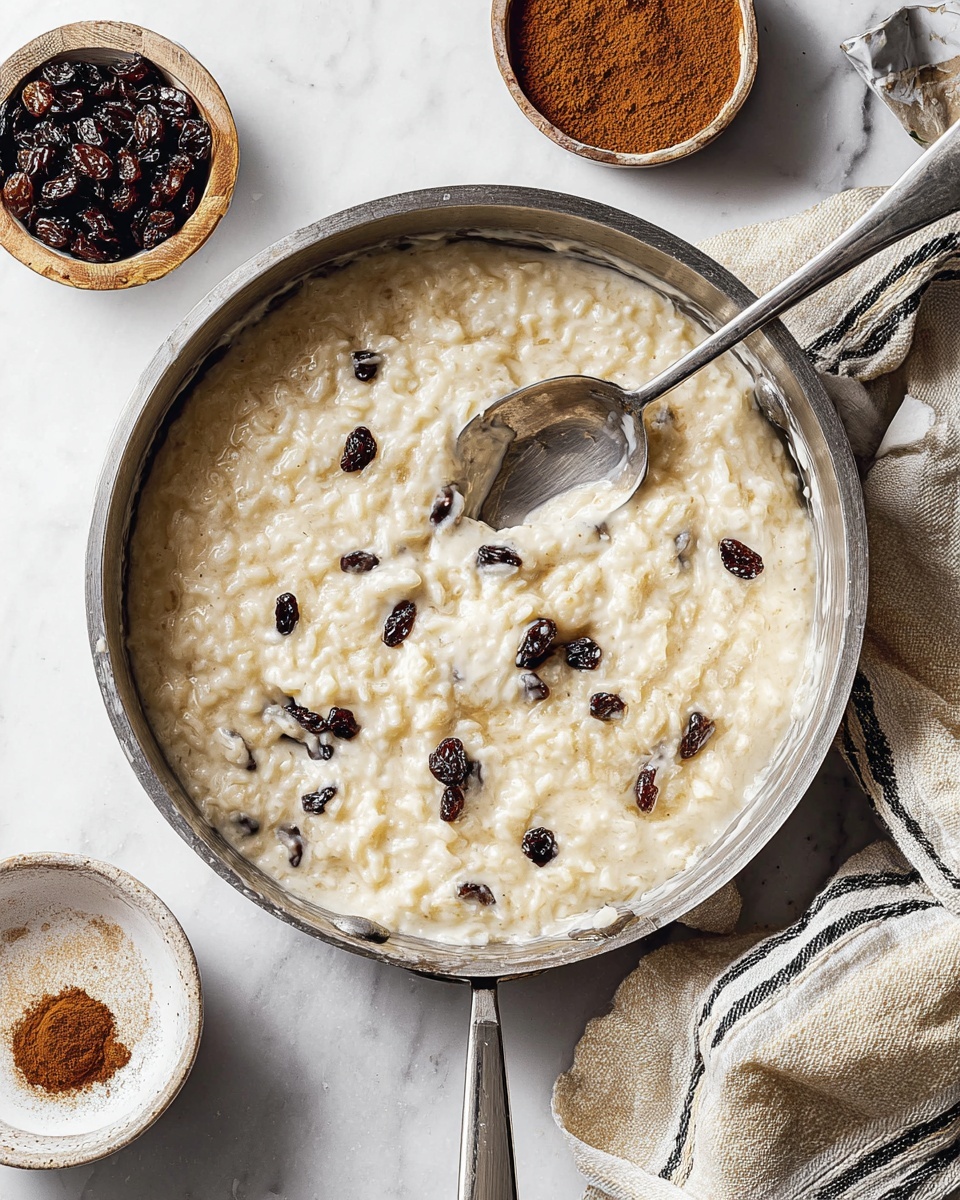 The image shows two bowls of creamy rice pudding placed on a white marbled surface. Each bowl has a thick layer of off-white rice pudding mixed with small dark raisins. On top of the pudding, a dusting of brown cinnamon powder is spread, along with a small cluster of dark raisins creating a warm contrast. One bowl holds a silver spoon resting inside, hinting at creaminess and softness. A white jar and a small white bowl with extra raisins sit nearby, while a striped white and beige cloth adds a soft texture to the scene. photo taken with an iphone --ar 4:5 --v 7