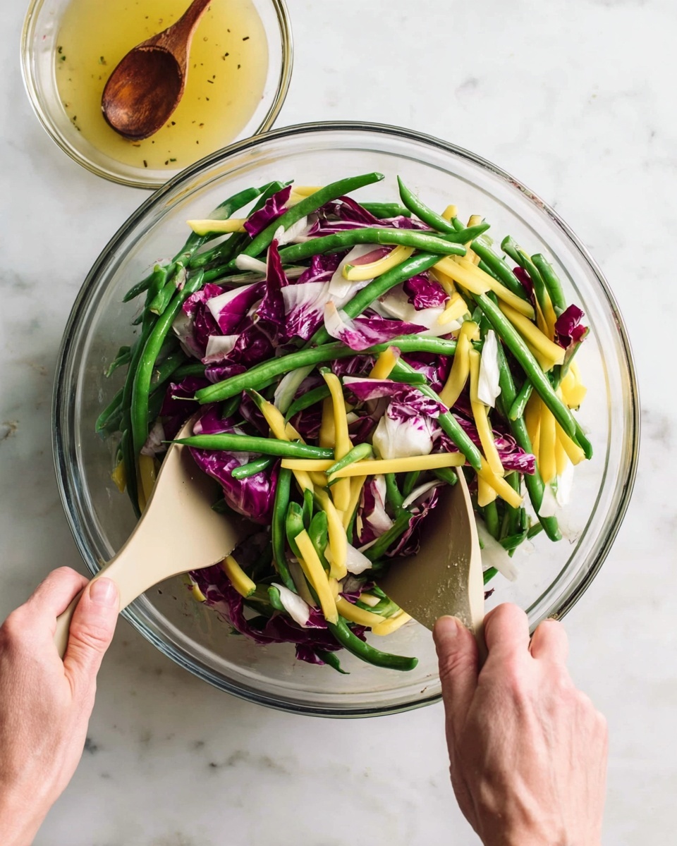 A clear glass bowl filled with a fresh salad showing vivid layers of green beans, yellow beans, and torn pieces of deep purple and white radicchio leaves mixed together. The green and yellow beans are long and smooth, creating a crisp texture, while the radicchio adds a rougher, leafy contrast scattered evenly throughout. Two woman's hands are holding beige salad utensils, one on each side of the bowl, tossing the salad gently. To the top left corner, there is a small clear glass bowl with a light yellow dressing and a wooden spoon resting inside. The whole scene is set on a white marbled surface. photo taken with an iphone --ar 4:5 --v 7