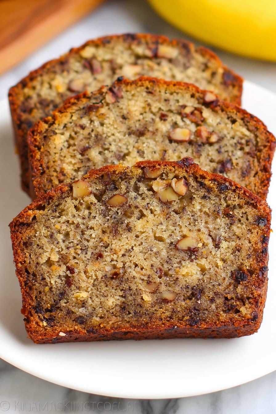 Three slices of banana nut bread are stacked slightly overlapping on a white plate, showing a dense texture filled with dark brown nuts and small chunks of banana inside the golden brown cake. The crust is a darker shade of brown and slightly crisp, while the inside looks moist with a mix of light and dark brown colors from the bananas and nuts. The plate is on a white marbled surface with a blurred yellow banana visible in the background. Photo taken with an iphone --ar 2:3 --v 7 - Moist Banana Bread with Walnuts and Raisins, banana bread recipes, easy banana bread, nutty banana bread, fruity banana bread