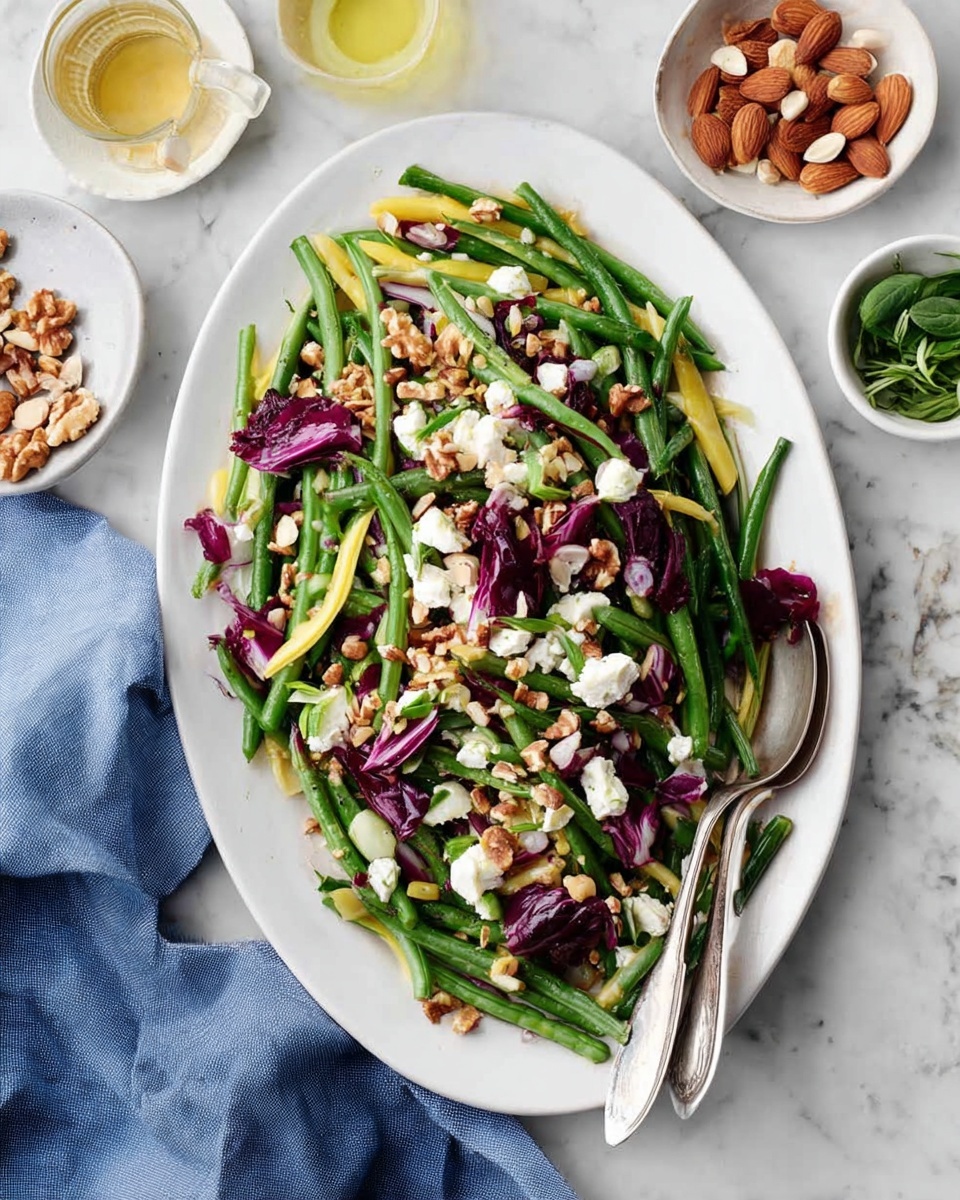 A white oval plate holds a fresh salad made of bright green beans and yellow beans as the first layer, mixed with deep purple-red radicchio pieces scattered on top. Soft white cheese chunks are spread evenly across the salad, with thinly sliced almonds and small walnut pieces sprinkled around for crunch. The salad rests on a white marbled surface with a silver fork and spoon placed on the plated side. Surrounding the plate are small white bowls containing more almonds and walnuts, a bottle of light yellow dressing, and a small bowl of green herbs. A folded blue cloth napkin is nearby. photo taken with an iphone --ar 4:5 --v 7