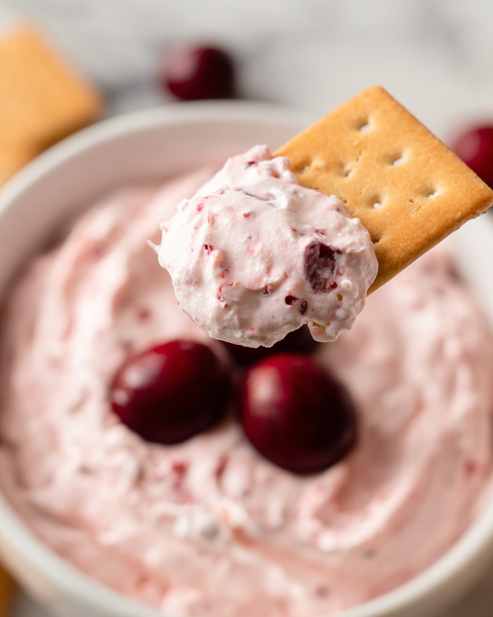 A close-up shows a light brown cracker dipped in a light pink creamy dip with small red berry pieces, held above a white bowl filled with the same pink dip with a smooth, swirled texture. Three dark red cherries rest on top of the dip in the bowl. The scene is set on a white marbled surface. photo taken with an iphone --ar 4:5 --v 7