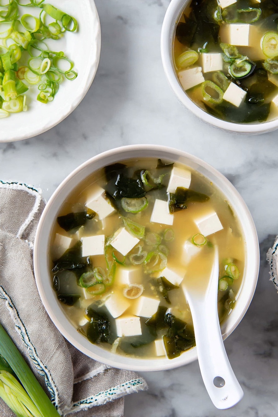 A white bowl filled with miso soup is shown from above, containing pale yellow broth with many small white tofu cubes, dark green seaweed pieces, and light green sliced scallions floating inside. A white spoon with a perforated handle is resting in the bowl, partly submerged in the soup. Nearby, a smaller white bowl matches the larger one and also holds the same soup with the same ingredients. A light gray cloth with white stitched edges lies near the bowls, and in the background, a white plate holds more sliced scallions. All items are placed on a white marbled surface. photo taken with an iphone --ar 2:3 --v 7 - Miso Soup with Tofu and Seaweed, easy miso soup recipe, healthy miso soup, tofu seaweed soup, quick Japanese soup