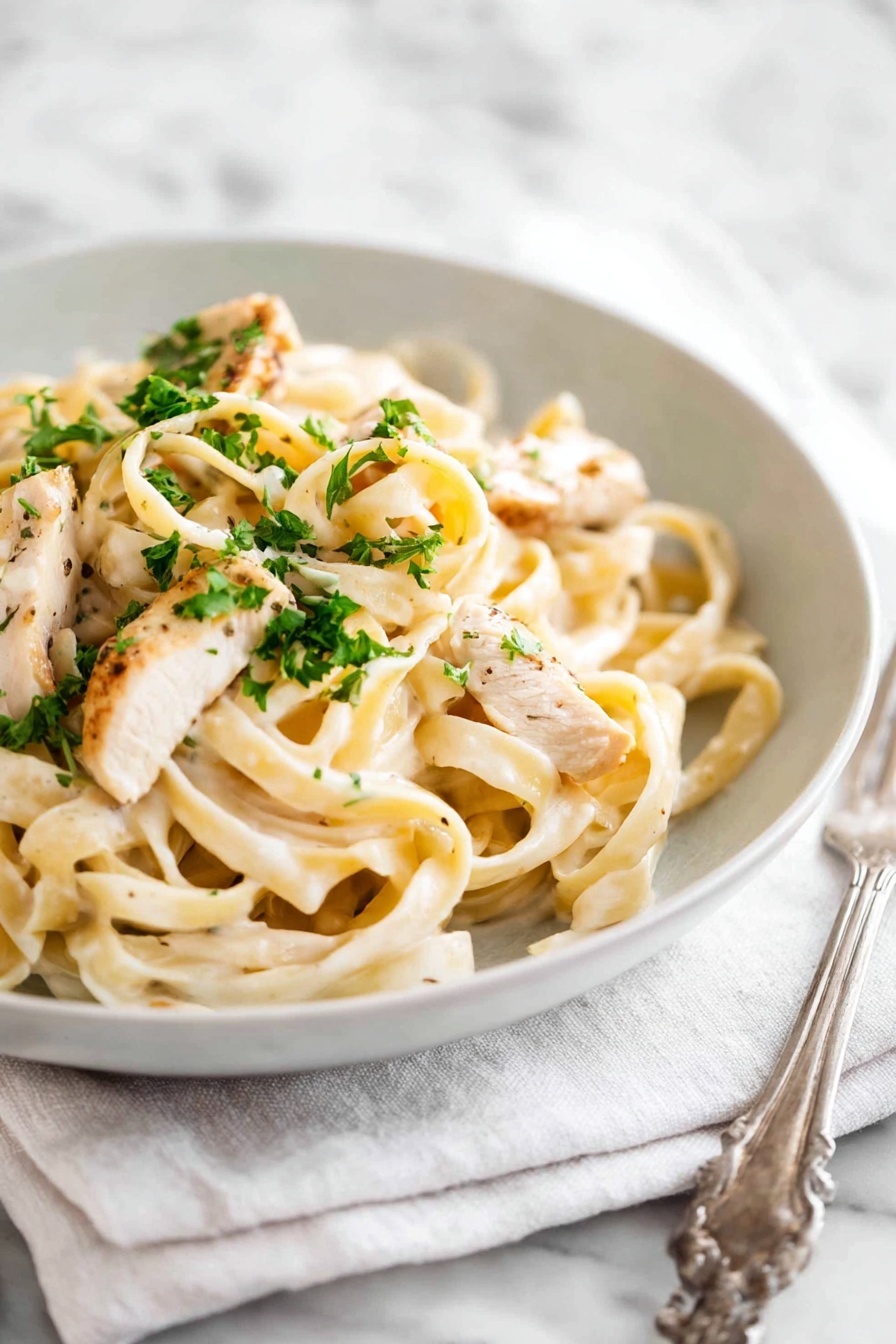 A white bowl filled with creamy fettuccine pasta topped with pieces of light golden-brown cooked chicken and sprinkled with small bright green parsley bits. The pasta is coated with a smooth, pale cream sauce and is twisted into soft loops. The bowl sits on a white cloth napkin, all placed on a white marbled surface. A silver fork is positioned to the left side of the bowl. Photo taken with an iphone --ar 2:3 --v 7 - One Pot Chicken Alfredo Pasta, creamy chicken pasta, easy Alfredo recipe, quick chicken pasta dinner, one pot creamy pasta