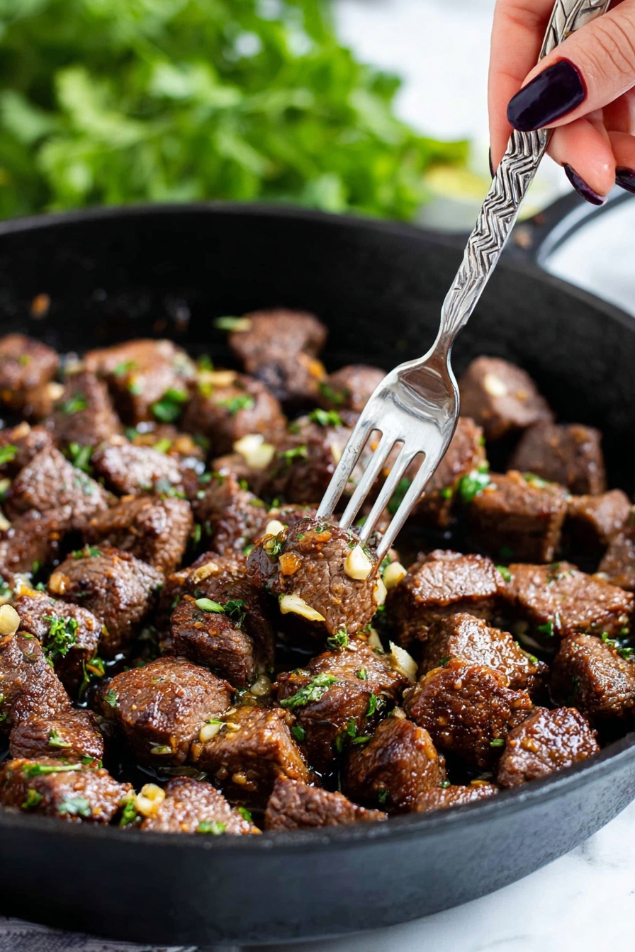 The image shows a black cast iron pan filled with many small chunks of cooked brown meat, each piece sprinkled with bits of green herbs and small pieces of golden-brown garlic. A woman's hand with dark nail polish is holding a silver fork that picks up a single piece of meat from the pan. The background is blurry with green leaves visible, creating a fresh feel. The pan sits on a white marbled surface. photo taken with an iphone --ar 2:3 --v 7 - Garlic Butter Steak Bites, garlic butter steak recipe, easy steak bites, juicy steak appetizers, quick steak dinner