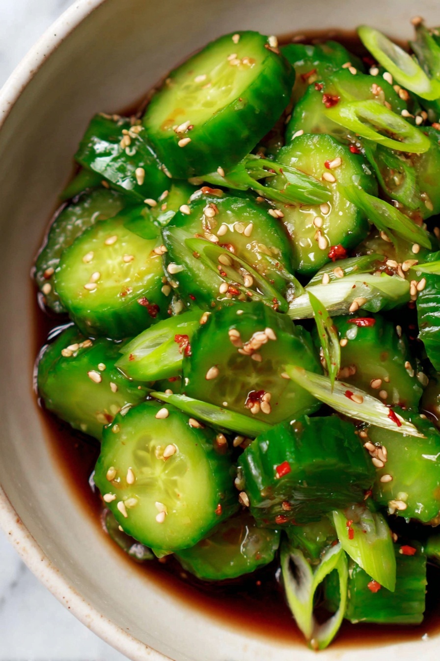 The image shows a close-up of a white bowl filled with bright green cucumber slices that have a slightly shiny, fresh texture. The cucumbers are cut into thick rounds and are mixed with thin slices of green onion. The dish is sprinkled with small, light tan sesame seeds and a few tiny red chili flakes scattered over the top. The cucumbers sit in a dark soy-based sauce that pools at the bottom of the bowl, and the overall look is vibrant and fresh against a white marbled surface. photo taken with an iphone --ar 2:3 --v 7 - Spicy Cucumber Salad, Spicy Cucumber Salad with Sesame and Garlic, Easy Summer Salad, Refreshing Cucumber Side Dish, Quick Healthy Salad